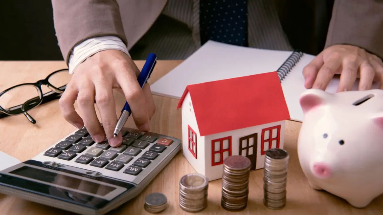 The image shows a person calculating home financing costs with a pen, surrounded by a toy house, stacks of coins, and a piggy bank, representing savings and budgeting for a home purchase.